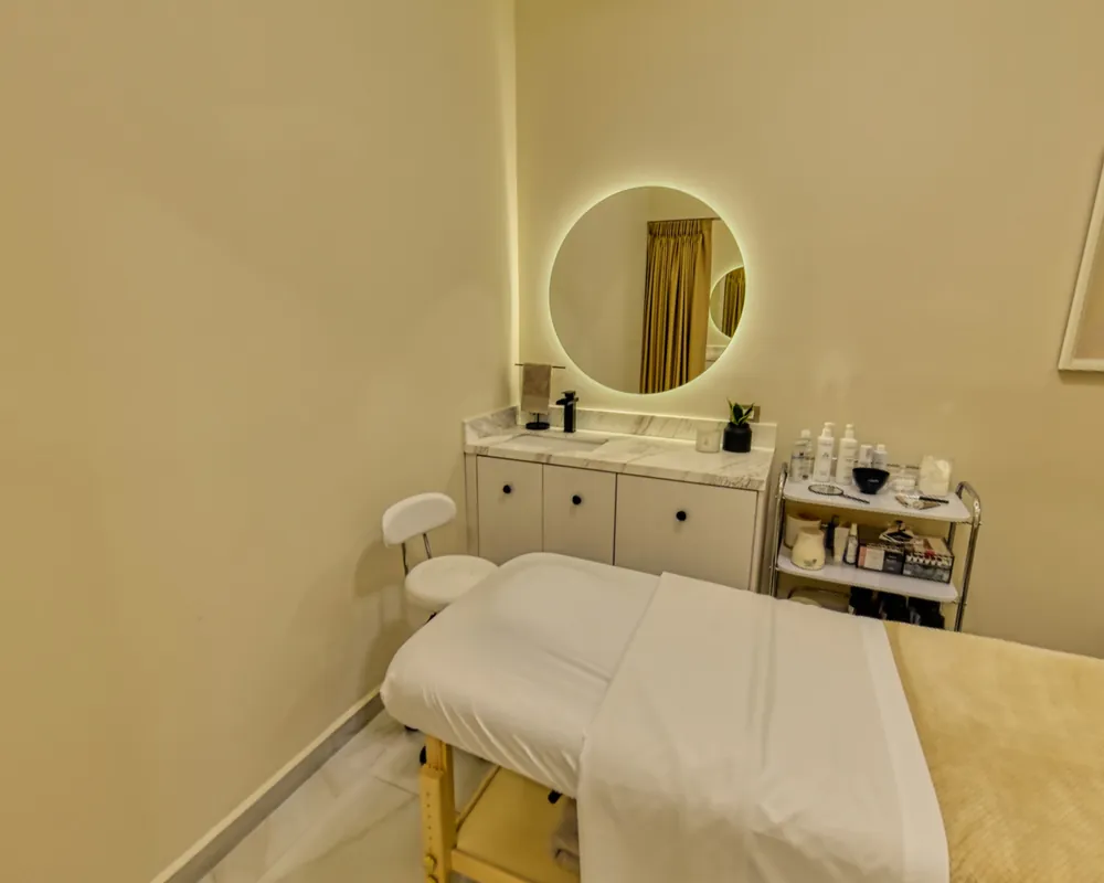 A facial room with a treatment bed, a marble vanity, a product trolley and a lit round mirror.