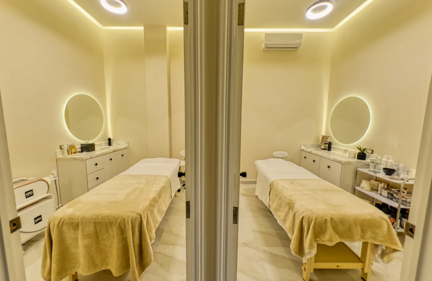Two open treatment rooms seen from the hallway, each with its bed, vanity and lit round mirror.